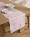 The Geometry Bubble Ditzy Dusty Rose stain-resistant table runner, featuring subtle purple stitching, lies on a wooden dining table topped with a stack of white ceramic plates.