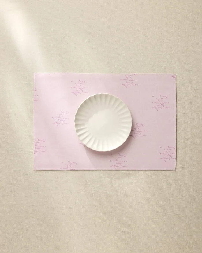 The Geometry Bubble Ditzy Dusty Rose placemat, featuring subtle lavender patterns, sits on a beige surface under soft lighting with a white scalloped-edge plate centered on top. Machine washable for easy care.