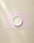 The Geometry Bubble Ditzy Dusty Rose placemat, featuring subtle lavender patterns, sits on a beige surface under soft lighting with a white scalloped-edge plate centered on top. Machine washable for easy care.