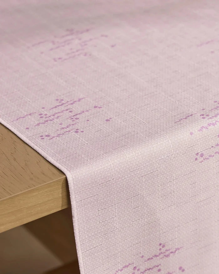 The Geometry Bubble Ditzy Dusty Rose table runner, featuring a light pink stain-resistant fabric with a subtle textured pattern and faint purple accents, drapes over the edge of a light wooden table.