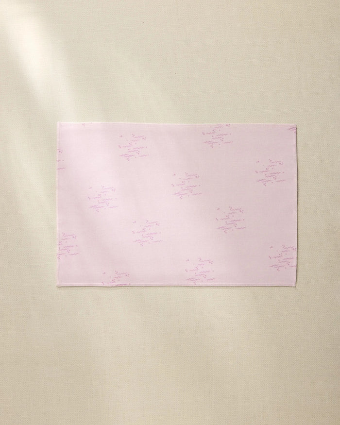 The Bubble Ditzy Dusty Rose placemat by Geometry is a rectangular pale pink cloth with subtle light purple star-like patterns, shown laid flat on a beige textured surface with soft lighting and a gentle left shadow.