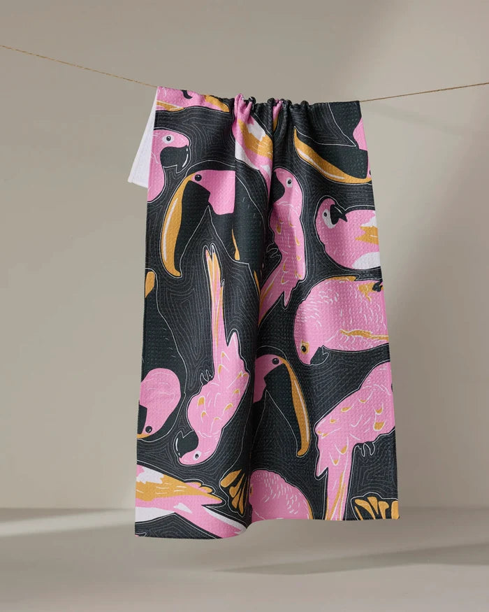 The Bubblegum Birds tea towel by Julianne Haness features bold pink birds with yellow and white accents on black, draped over a line against a neutral backdrop. Crafted from ultra-absorbent recycled materials.