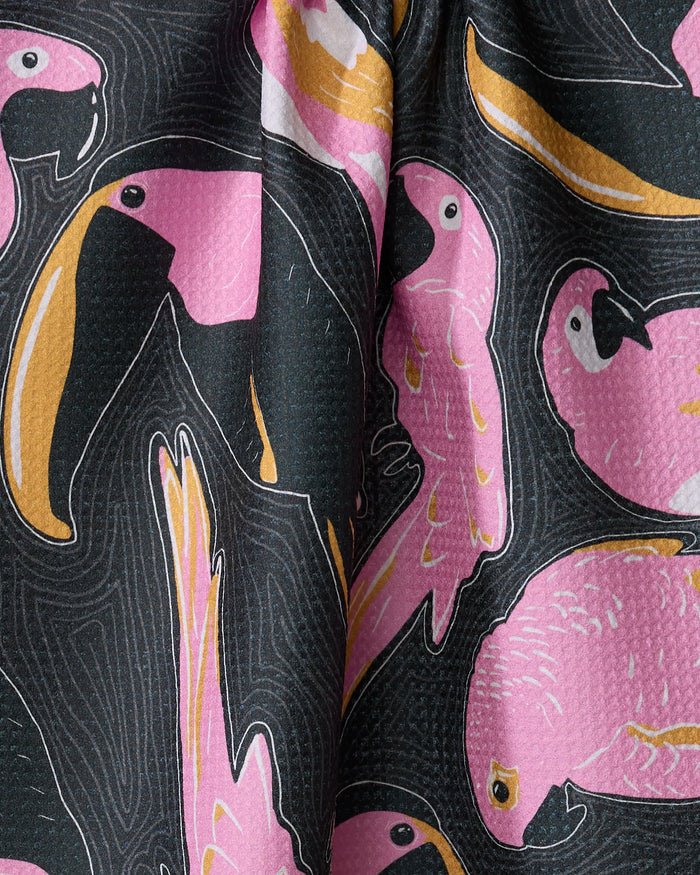 The Bubblegum Birds tea towel by Julianne Haness features playful illustrated toucans and parrots in pink, yellow, and black tones on a dark background, made from slightly creased recycled materials.