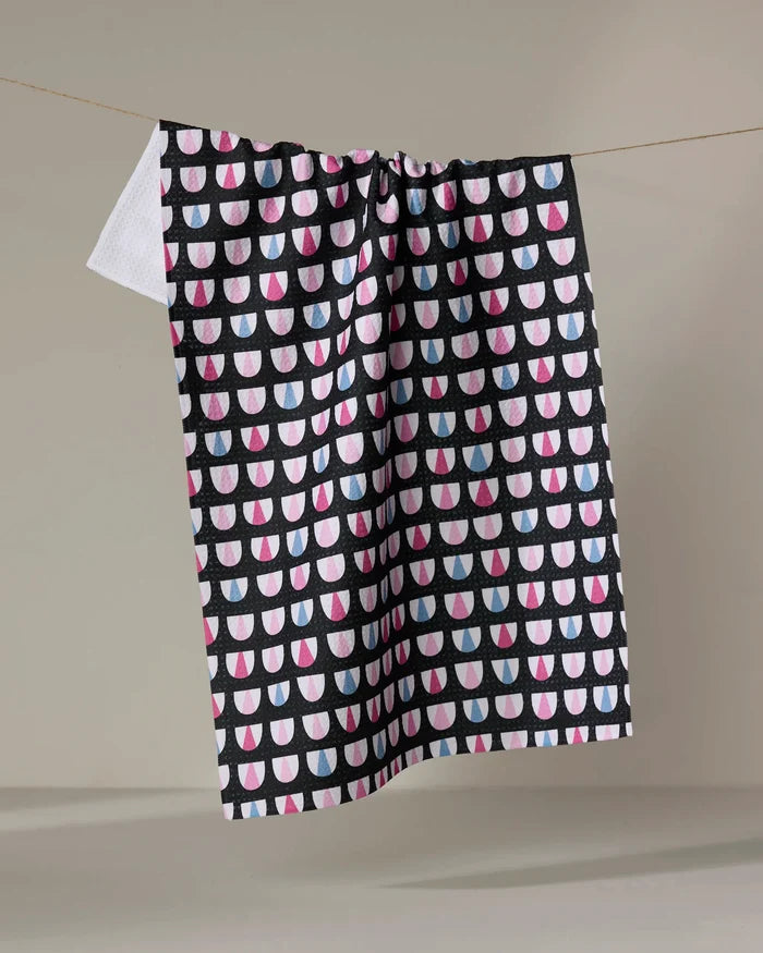 The Bug Boogie recycled kitchen towel by Anette Heiberg, with a geometric pattern of pink, white, and blue semi-circles on black, hangs on a line against a plain beige backdrop.