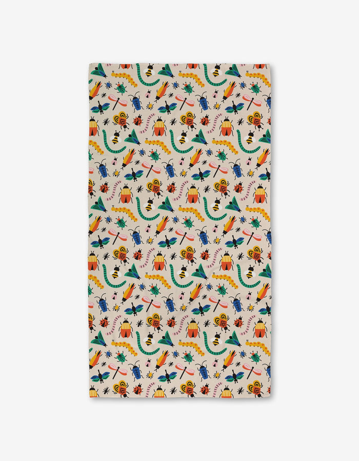 A rectangular cloth from Genna Blackburn called Bugs, made with eco-friendly towels, features a playful pattern of bugs in green, yellow, red, and blue with volcanoes, trees, and scattered leaves on a white background.