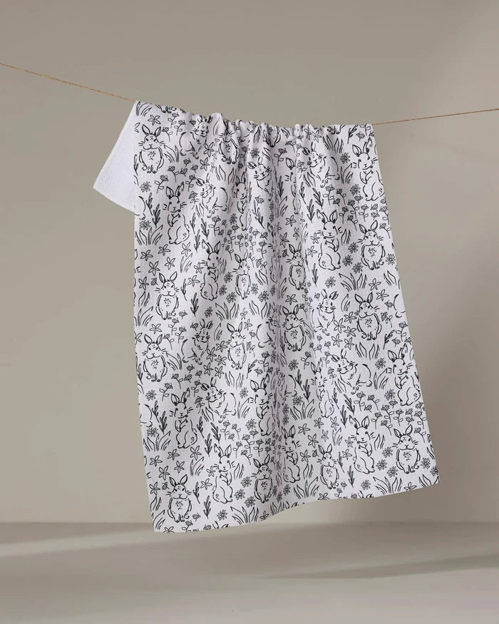 The Bunny Rabbit Garden towel by Charlie Rowan Designs hangs from a clothesline, its charming black floral pattern standing out against a simple, neutral background.