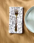 A gold fork rests on a Charlie Rowan Designs Bunny Rabbit Garden Geosuede dinner napkin set, featuring black bunny and floral illustrations, beside a light blue plate on a wooden table.