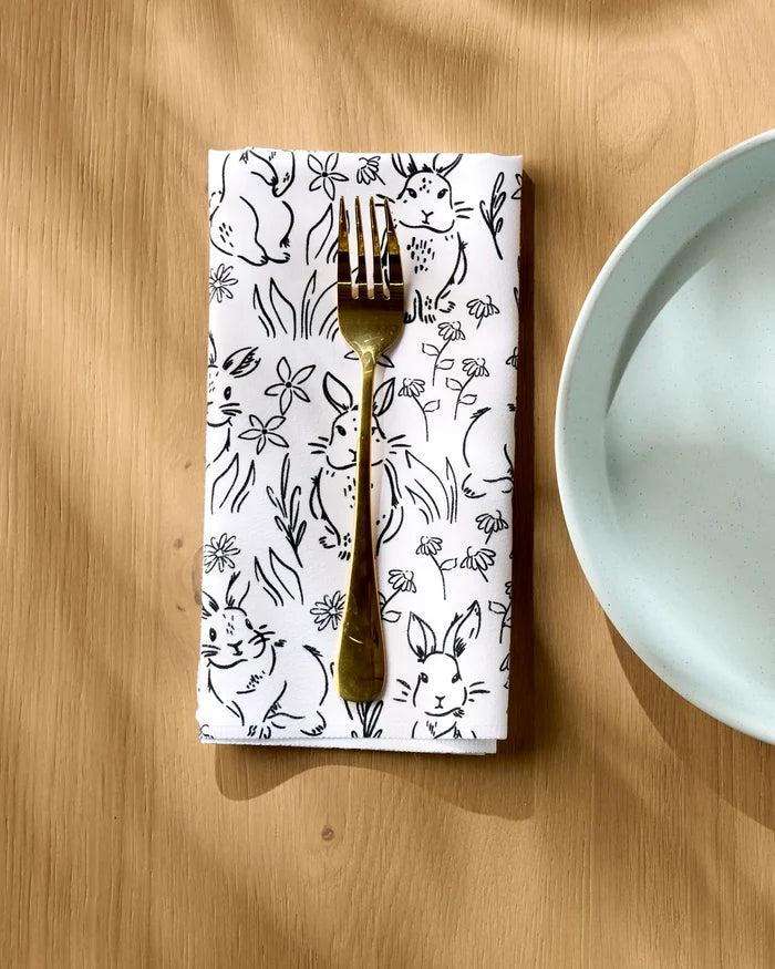 A gold fork rests on a Charlie Rowan Designs Bunny Rabbit Garden Geosuede dinner napkin set, featuring black bunny and floral illustrations, beside a light blue plate on a wooden table.