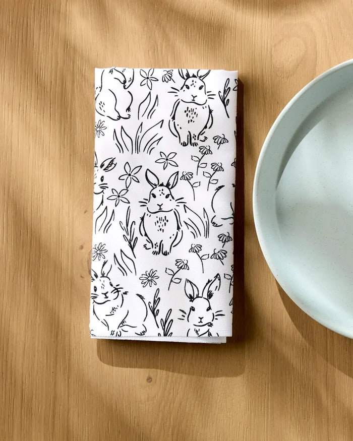 A "Bunny Rabbit Garden" recycled dinner napkin by Charlie Rowan Designs, featuring black bunny and flower sketches, is folded on a light wooden table next to a light blue plate.