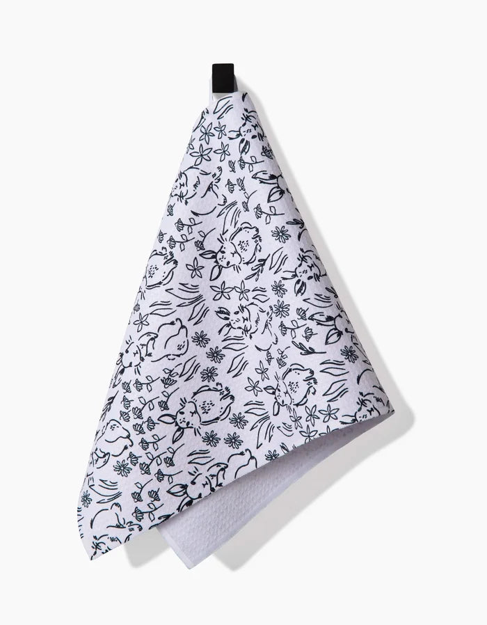 The Bunny Rabbit Garden kitchen towel by Charlie Rowan Designs features black floral and botanical line drawings on white recycled fabric, hanging from a black loop against a white background.