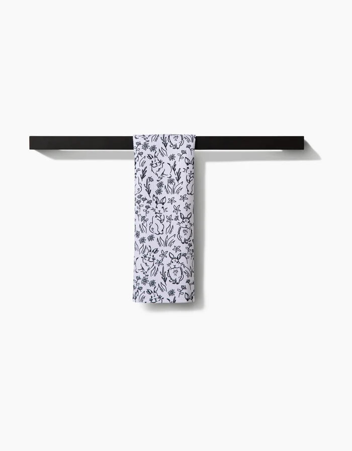 The Bunny Rabbit Garden towel by Charlie Rowan Designs, featuring a black floral pattern, hangs neatly folded over a sleek black towel bar against a plain white background.
