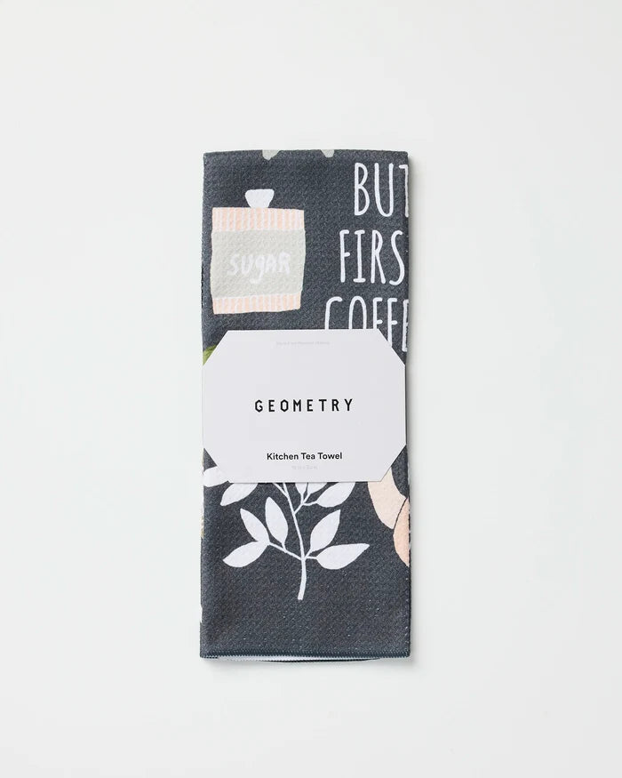 A folded tea towel made from recycled materials featuring a plant and text design, wrapped in a white label that reads "But First, Coffee" by Cottonflower Studio, placed on a plain white background.