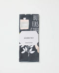 A folded tea towel made from recycled materials featuring a plant and text design, wrapped in a white label that reads "But First, Coffee" by Cottonflower Studio, placed on a plain white background.