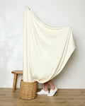 A person stands on a wooden floor, mostly covered by Geometry’s large Butter Yellow blanket. Only their lower legs and white sneakers are visible. Nearby sit a woven basket and a small wooden stool against a plain wall.
