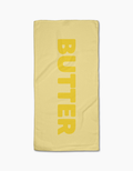 The GEOMETRY Butter Bar Towel is pale yellow, made from Geoweave fabric, and features BUTTER in bold vertical yellow letters on a textured surface. Displayed on a white background.