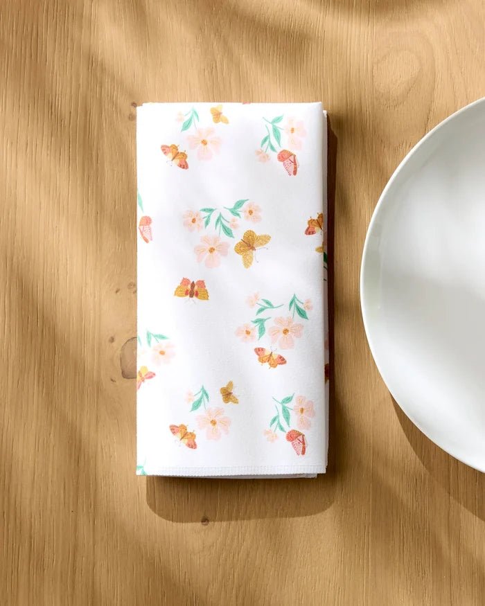 The "Butterfly Garden" cloth napkin by Danelys Sidron, featuring a floral and butterfly print, is neatly folded on a wooden table beside a white plate, with sunlight casting gentle shadows over the scene.