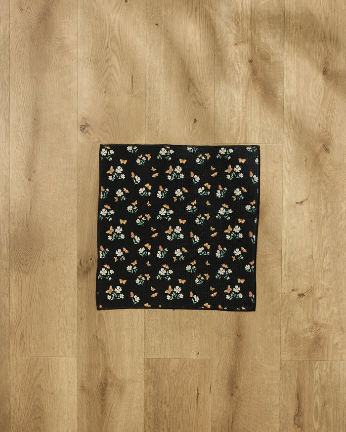 The Danelys Sidron Butterfly Garden towel, small and black with a vibrant floral design, is laid flat on a light brown wooden floor.