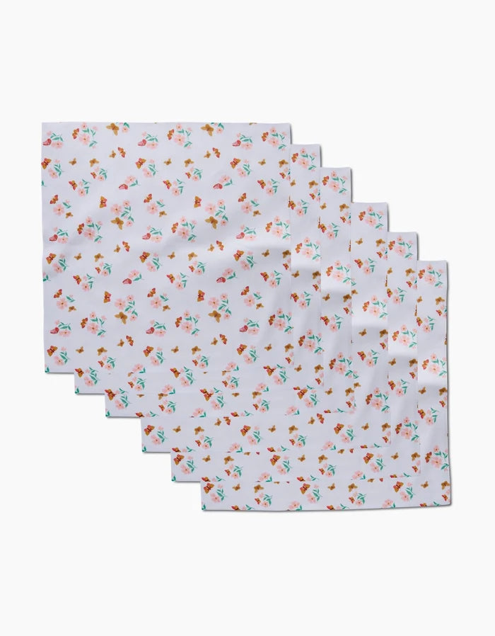 Six white Butterfly Garden Dinner Napkins by Danelys Sidron, made from recycled materials and featuring a small pink, orange, and green floral pattern, are arranged in a staggered row on a plain background.