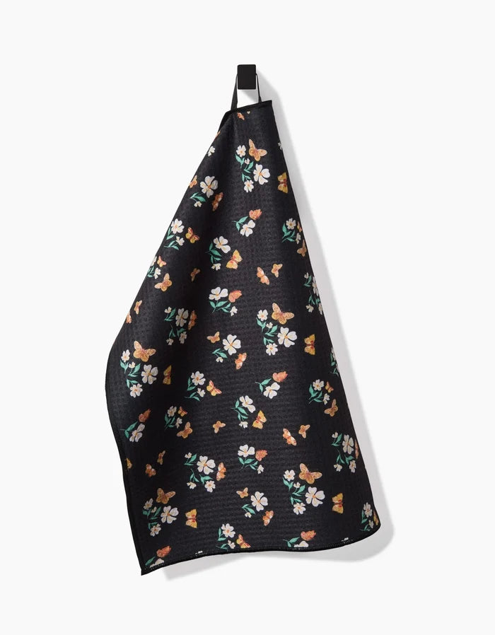 The Butterfly Garden dish towel by Danelys Sidron features ultra-absorbent Geoweave fabric with a vibrant floral and butterfly pattern, shown hanging from a black loop on a plain white background.