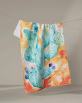 The Skye Walker "Cacti" tea towel showcases a quick-drying design with a vibrant watercolor green cactus and purple flowers on a beige background.