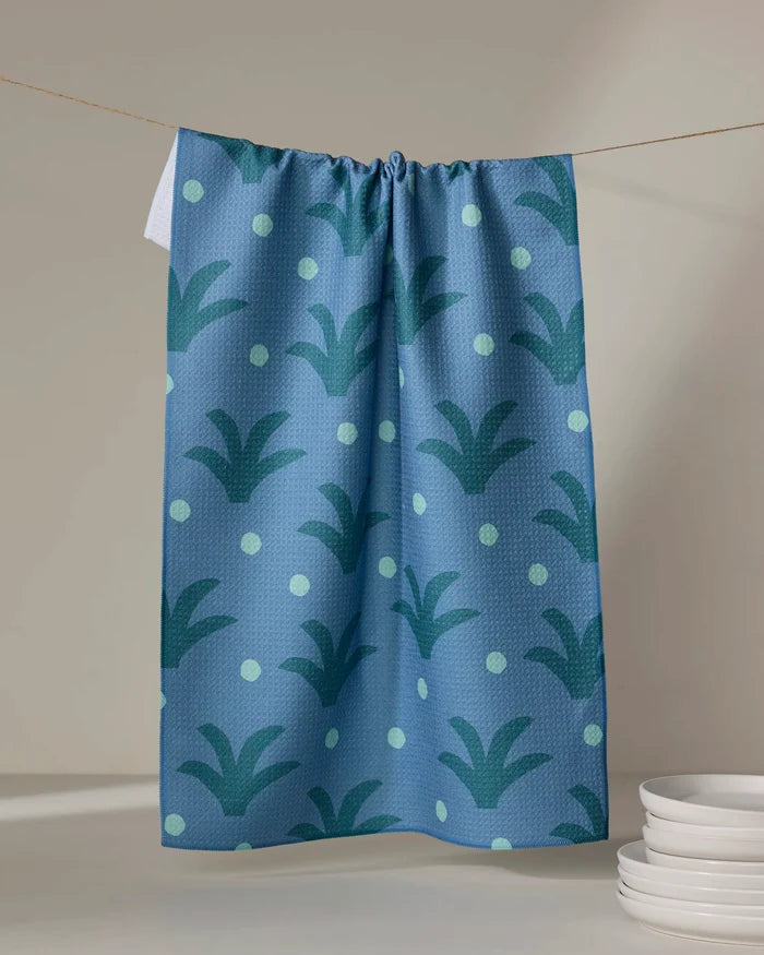 The Elizabeth Edwards Cactus Dot tea towel features blue fabric with green polka dots and abstract leaf patterns. Hanging on a clothesline, it’s made from ultra-absorbent recycled materials and pairs beautifully with stacked white plates.