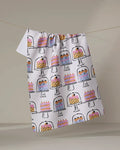 The "Cakes In The Window" tea towel by Megan Roy features vibrant cake illustrations on stands, crafted from recycled materials and displayed hanging against a plain background.