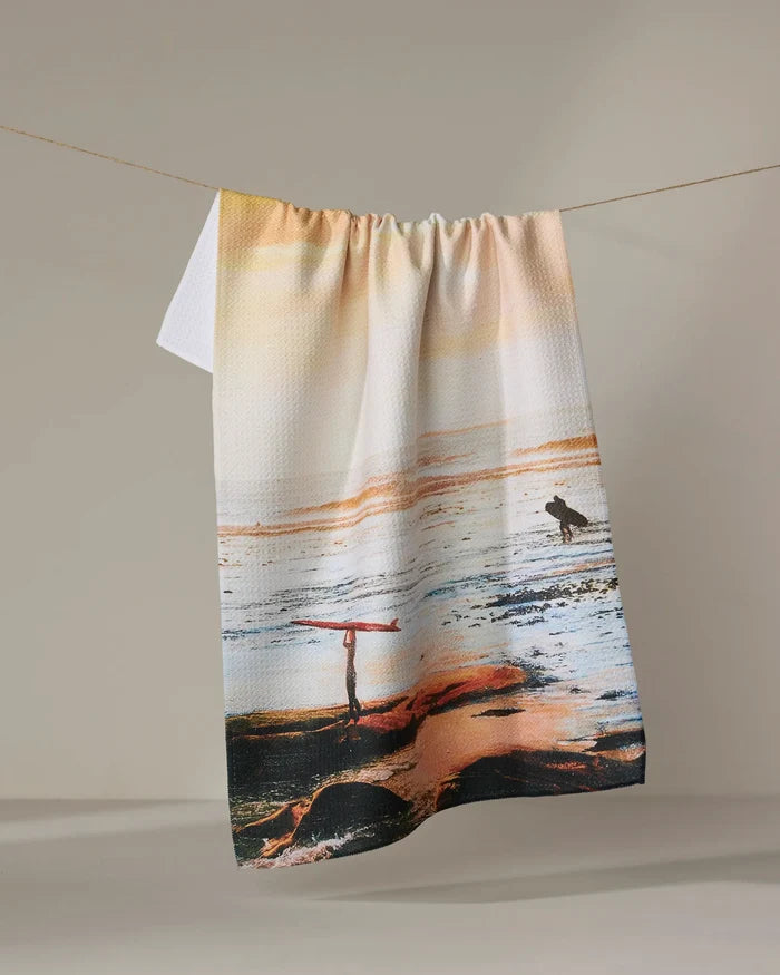 The Call Of The Wild tea towel by Stefanie Bales features two surfers on a pastel beach scene, made from ultra-absorbent, recycled materials.