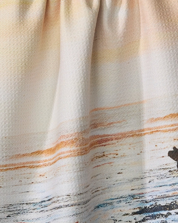 A close-up of the Stefanie Bales "Call Of The Wild" tea towel shows fabric gathered at the top, featuring an ultra-absorbent material and an ocean sunset design in orange, yellow, and blue over water.