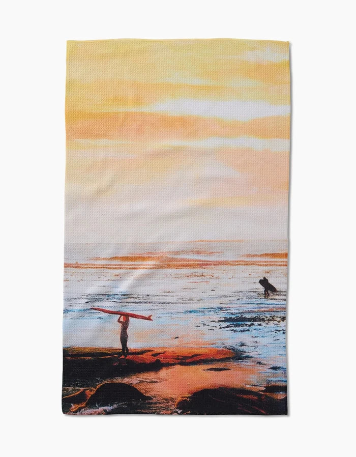 The Call Of The Wild tea towel by Stefanie Bales is made from recycled materials and features two surfers at sunset—one on shore with a board, one in the water—against vibrant orange and yellow sky reflections.