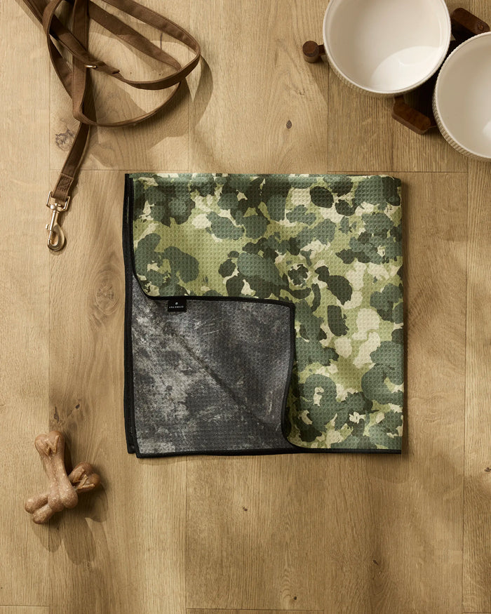 Geometry’s Camo towel, a green and gray camouflage dog towel made from recycled materials, sits on a wooden floor beside two dog bowls on a stand, a brown leash with metal clasp, and two bone-shaped toys.