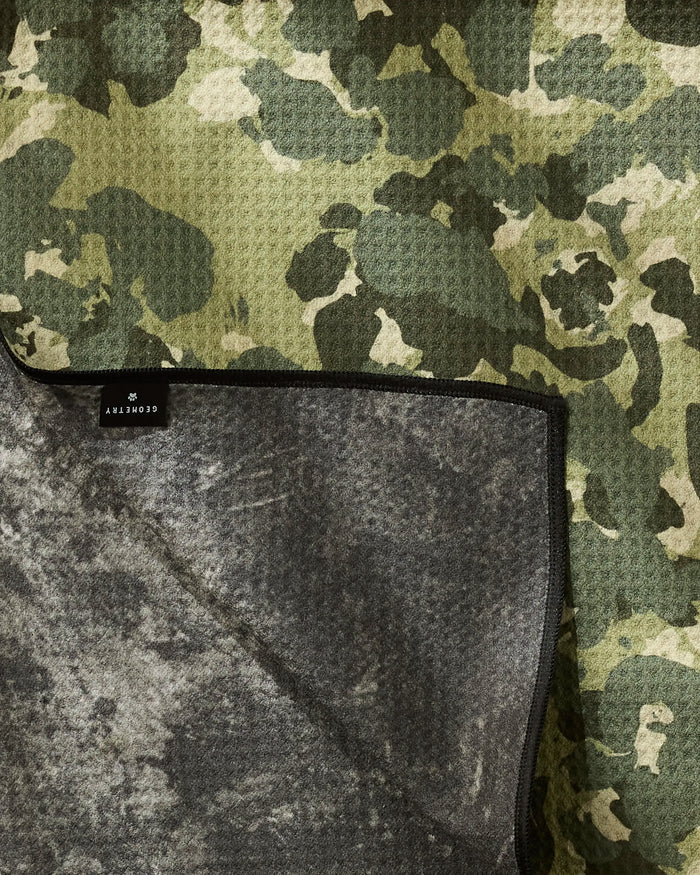 A close-up of Geometry’s Camo dog towel, featuring a green and beige camo pattern, textured gray underside, folded corner, and a small black label with white text. Made from recycled materials.