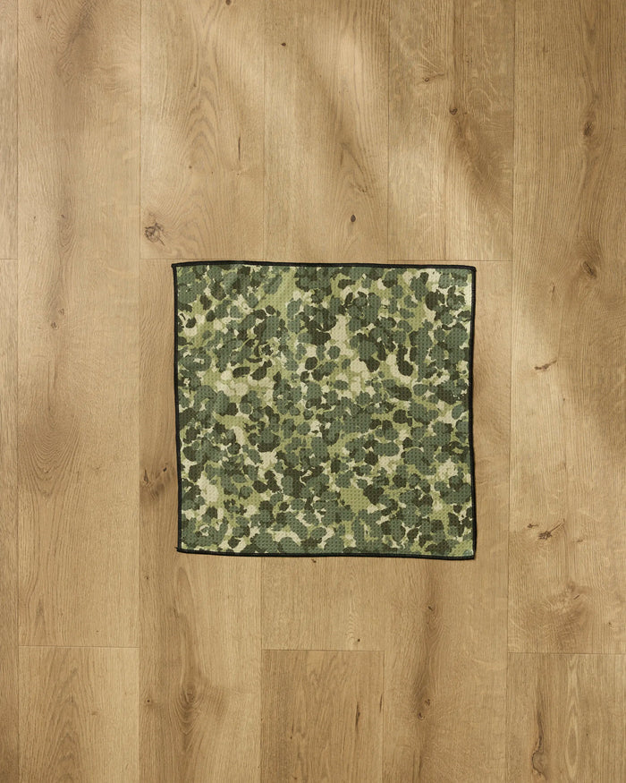 A small rectangular Geometry Camo paw towel with a green camouflage pattern is placed on a light brown wooden floor.