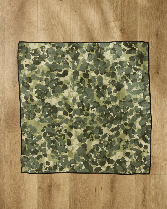The Geometry Camo square dog towel, made from recycled materials, features a green and brown camouflage pattern with a black border and is shown laid flat on a light wooden floor.