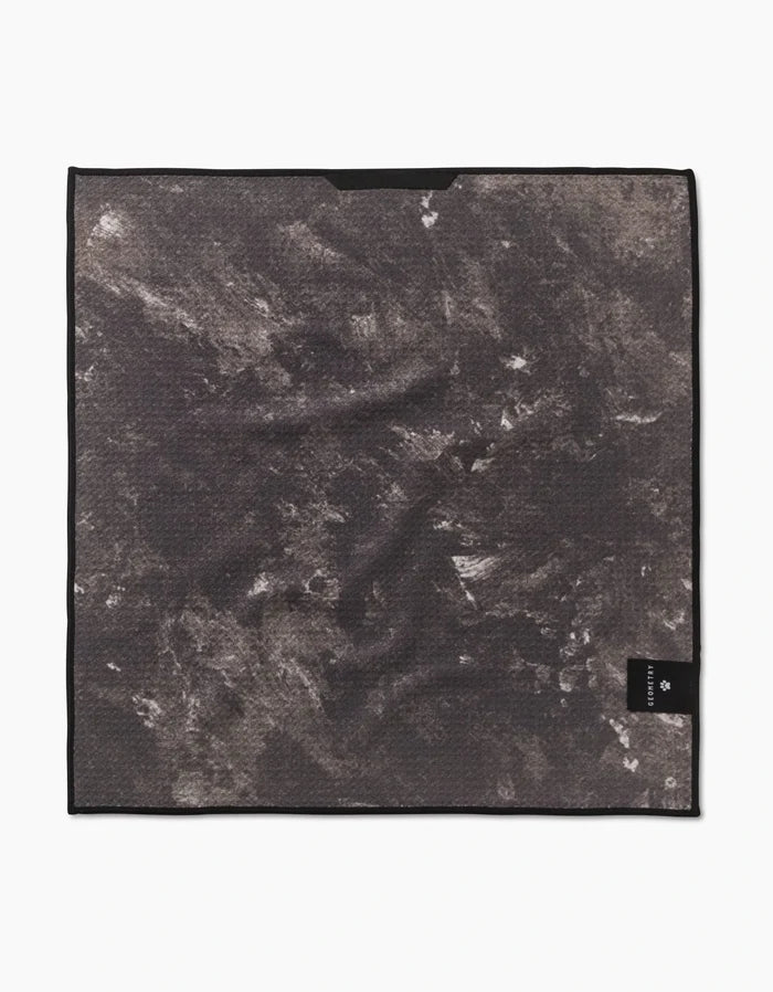 The Geometry Camo Paw Towel is a square, ultra-absorbent Geoweave towel in a textured black and gray abstract pattern. It has a black border and features a small black tag with white text in the bottom right corner.