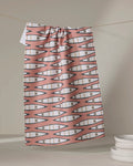 A "Canoes" tea towel by Coit Creative, featuring a pink and white geometric zigzag design and made from quick-drying recycled materials, hangs on a string near stacked white plates against a neutral backdrop.