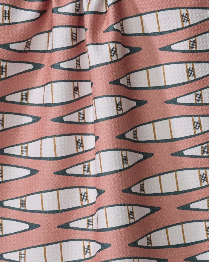 The Coit Creative "Canoes" tea towel features a repeating pattern of white canoes outlined in black and tan on a muted pink background. Made from recycled materials, the fabric includes subtle folds and gentle shadows.