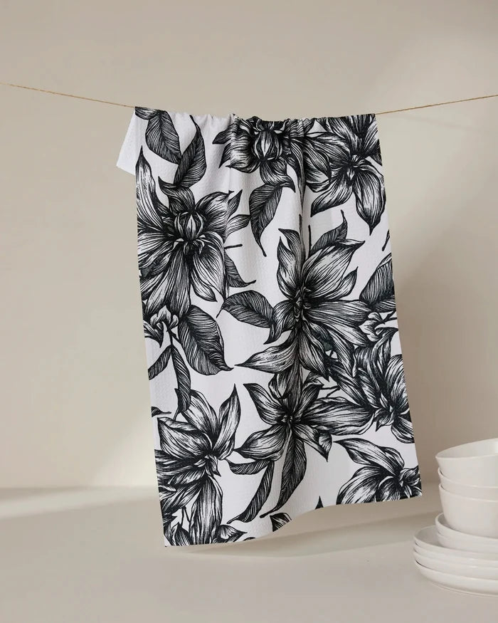 The GEOMETRY Caring tea towel, featuring a bold black floral pattern on ultra-absorbent white fabric, hangs from a string beside stacked white bowls and plates on a neutral background.
