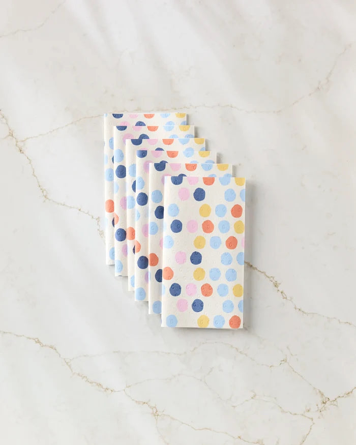 Six Carnival kitchen towels by Lizzie Clark, featuring eco-friendly fabric and blue, orange, pink, and purple polka dots, are stacked in a staggered row on a white marble surface.