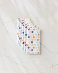 Six Carnival kitchen towels by Lizzie Clark, featuring eco-friendly fabric and blue, orange, pink, and purple polka dots, are stacked in a staggered row on a white marble surface.