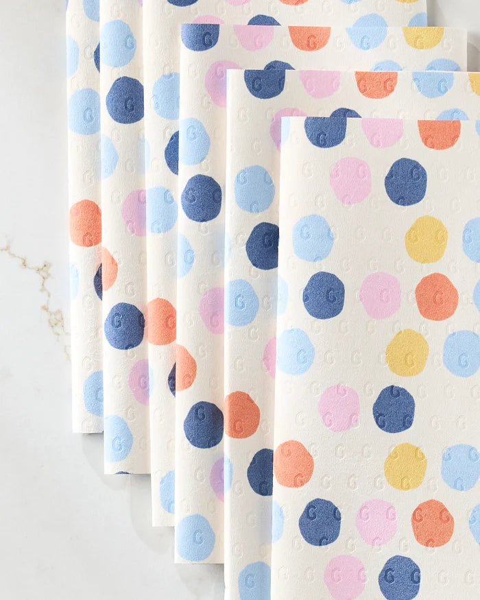 Lizzie Clark's Carnival eco-friendly kitchen towels, featuring colorful blue, pink, orange, and yellow polka dots, are displayed fanned out on a light marble surface.