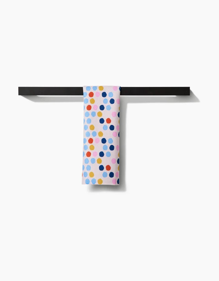 The Carnival bar towel by Lizzie Clark, crafted from recycled materials, features colorful blue, yellow, orange, and pink polka dots on white and hangs neatly over a simple black bar against a white background.