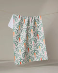 The "Carrot Damask" dish towel by Sarah Daugherty, featuring an illustrated orange carrot and green leaf pattern, hangs on a clothesline against a neutral background.