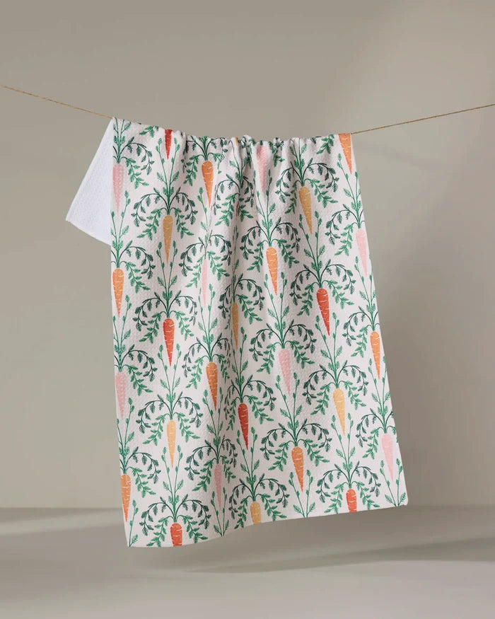 The "Carrot Damask" dish towel by Sarah Daugherty, featuring an illustrated orange carrot and green leaf pattern, hangs on a clothesline against a neutral background.