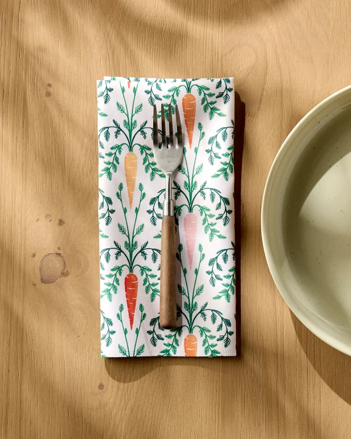 A Sarah Daugherty Carrot Damask recycled napkin with carrot and leaf designs holds a wooden-handled fork beside a ceramic bowl on a wooden table.