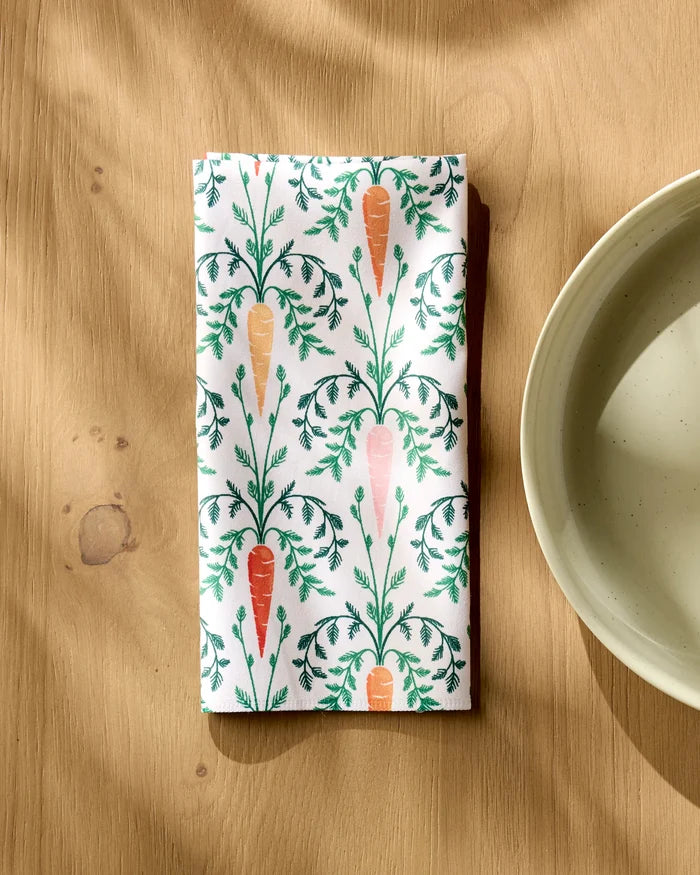 A folded Carrot Damask dinner napkin by Sarah Daugherty, featuring a carrot and green leaf pattern on soft Geosuede, rests on a light wooden table beside a beige ceramic bowl as sunlight casts gentle shadows.