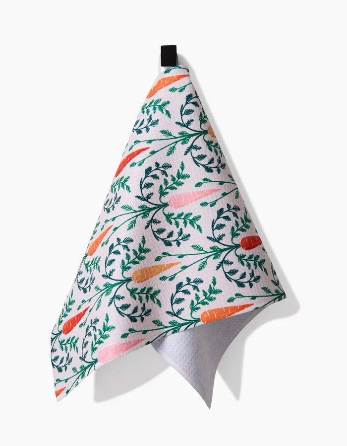 The Sarah Daugherty Carrot Damask recycled kitchen towel, featuring a colorful carrot and green leaf pattern, hangs from a black loop against a plain white background.