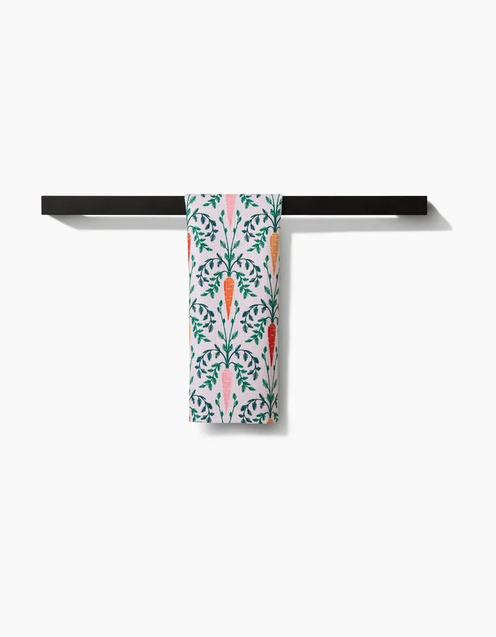 The Sarah Daugherty Carrot Damask recycled kitchen towel, featuring vivid orange carrots and green leaves, hangs neatly on a black bar against a plain white background.