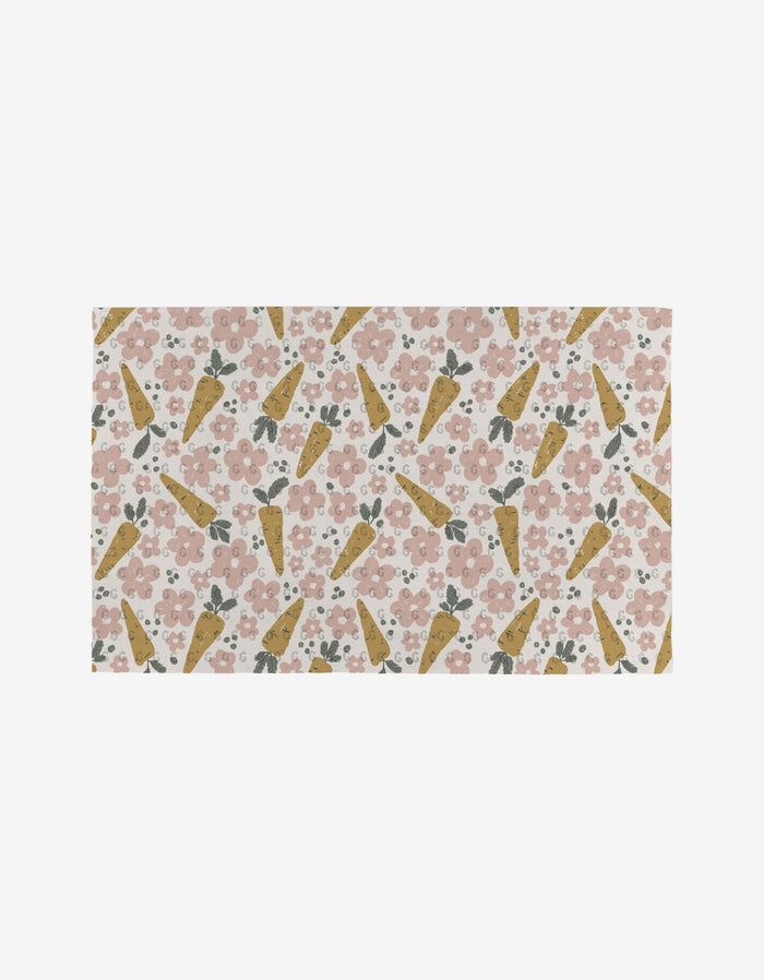 The Carrot Patch mat by Charlie Rowan Designs is a rectangular, eco-friendly mat with a repeating pattern of yellow carrots, pink flowers, and green leaves on a light background.