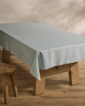 A wooden table draped with the Geometry Chambray Light Blue sustainable tablecloth stands on a wooden floor beside a small wooden stool, with a softly blurred neutral-toned background.
