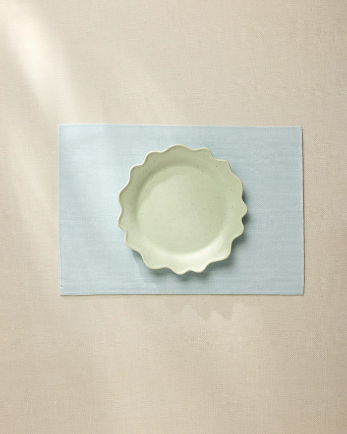 A light green, wavy-edged ceramic bowl sits centered on the Geometry Chambray Light Blue placemat, which rests on a beige tablecloth under soft, natural light.